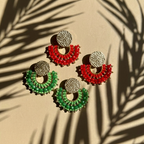 Pair of red and green earrings on a beige surface with leaf shadows.