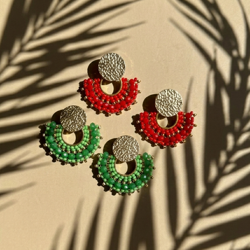 Pair of red and green earrings on a beige surface with leaf shadows.