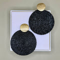 Black circular earrings with gold accents on a white box against a beige background