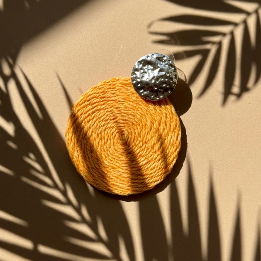 Round woven earring with a metallic knob on a beige surface with palm leaf shadows