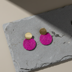Pair of magenta textile earrings with gold round tops on a gray stone surface.