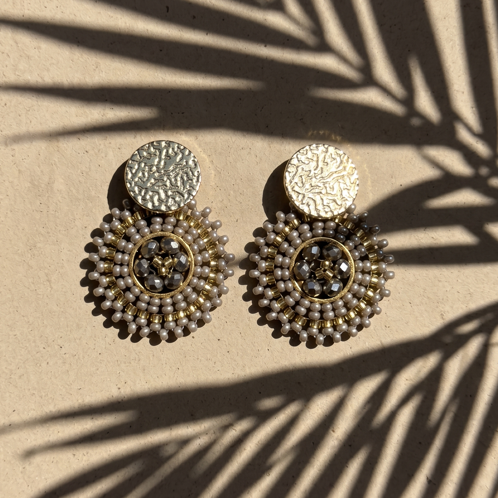 Pair of grey beaded earrings on a beige surface with palm leaf shadow