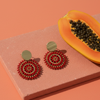Red beaded earrings on a pink stand with a halved papaya on an orange background