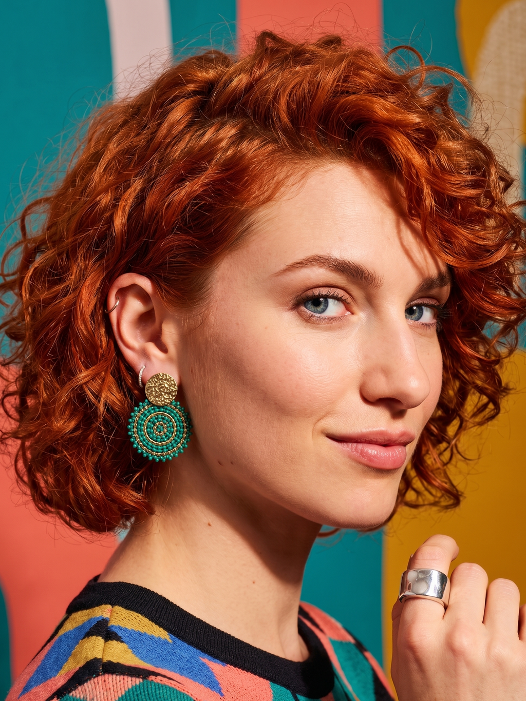 Woman with red hair wearing colorful earrings and a multicolored top against a multicolored background